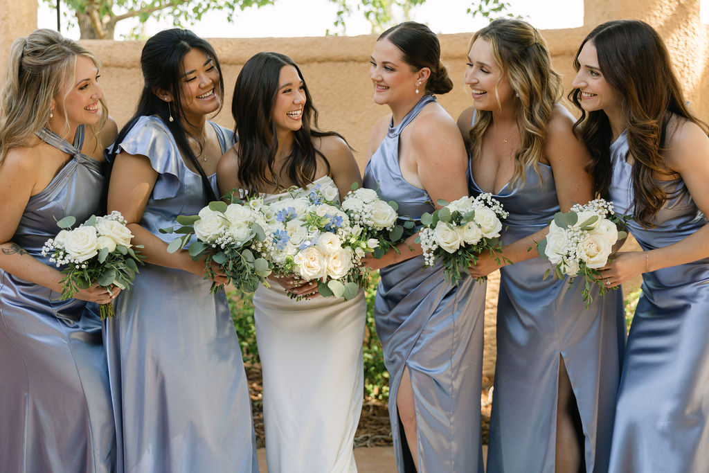 Bridesmaids laughing with the bride while holding white and blue bouquets during a Villa Parker garden wedding in Colorado.