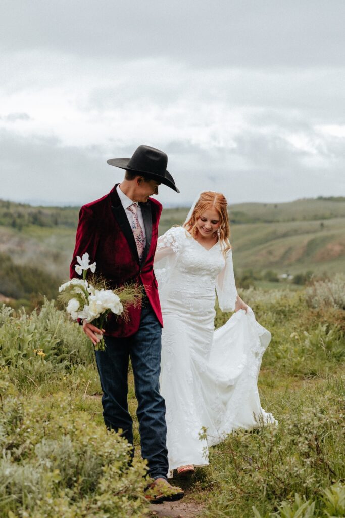 Groom candidly leading his bride through the mountains while carrying her white bridal bouquet