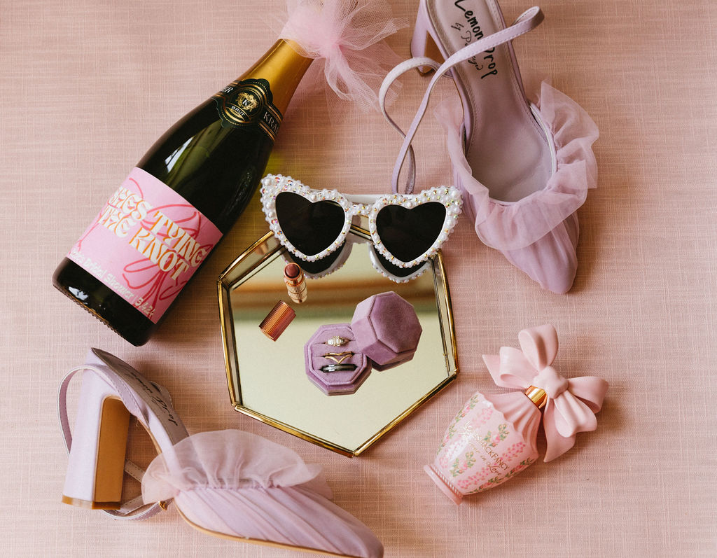 Colorful, pink bridal details arranged with champagne, lipstick, perfume, and heart sunglasses at a surprise elopement. 