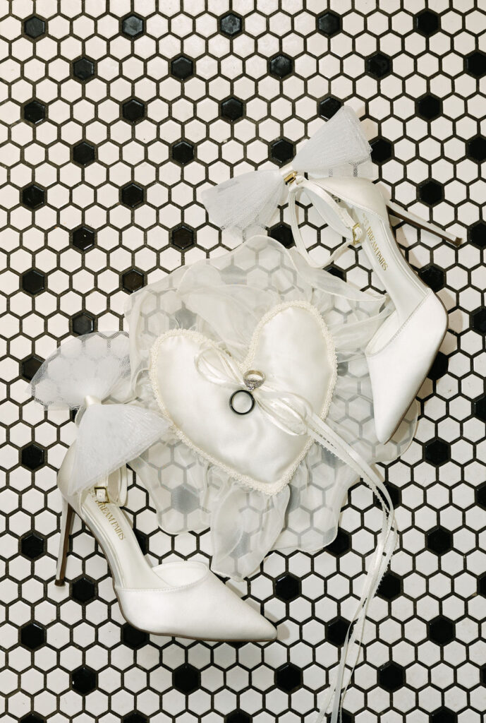 Vintage wedding details with rings styled on a vintage heart pillow with lace on a checkered tiled floor. 