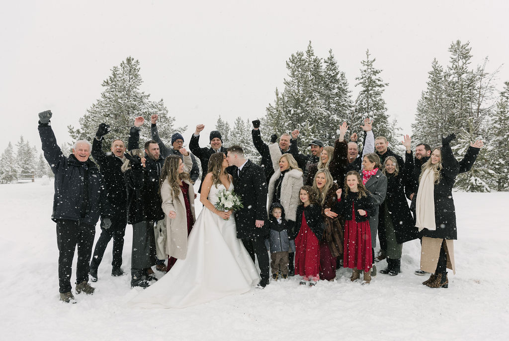 Outdoor snowy winter wedding joyful family photo with bride and groom kissing