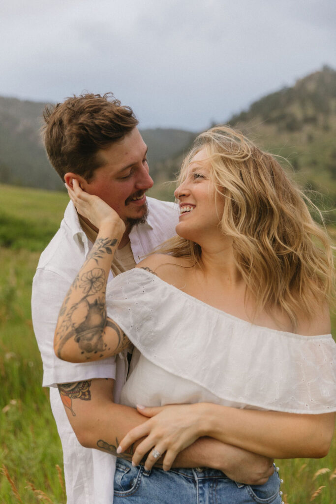 Intimate close-up of Paige holding Austin's face while laughing together, capturing authentic joy during their Colorado wildflower engagement session at Chautauqua Park