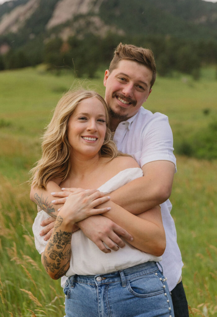 Joyful portrait of couple hugging as they both smile at camera with a stunning field and Flatirons mountains in background