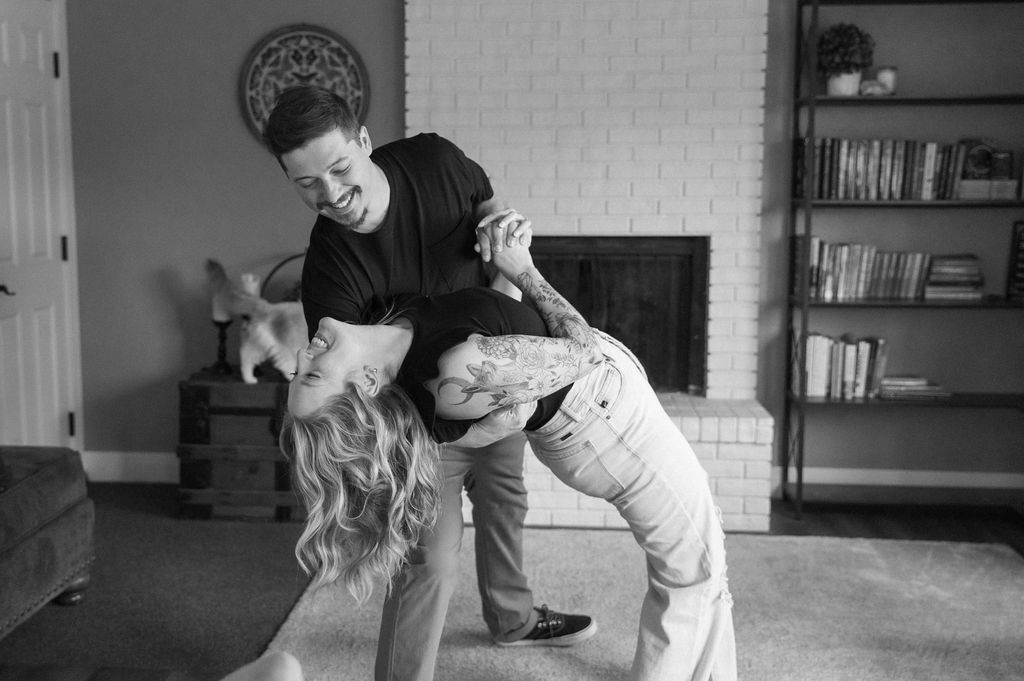 Black and white photo of couple dancing in their living room with their cat in the background during a natural engagement session. 