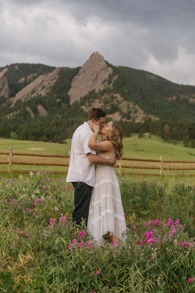 Paige and Austin embracing while surrounded by pink Colorado wildflowers with dramatic mountain backdrop at Chautauqua Park