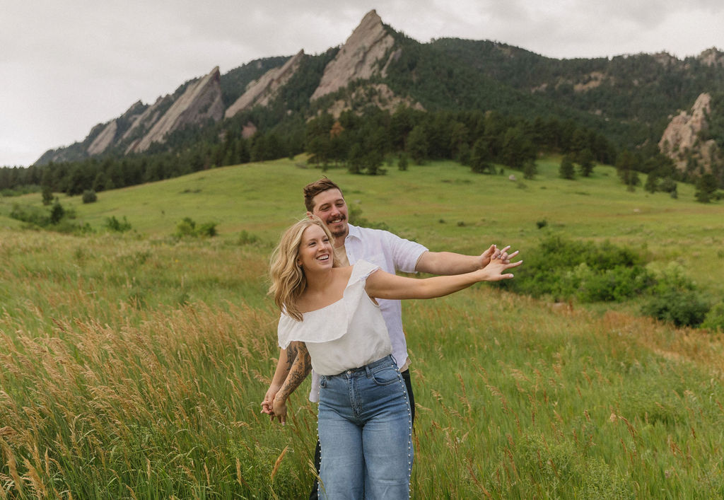 Laughing engaged couple dancing in field of pink wildflowers during golden hour at Chautauqua Park in Boulder