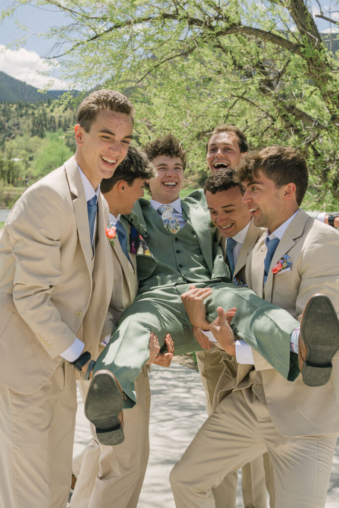 Groomsmen in coordinating suits laughing together at outdoor wedding with mountain forest background