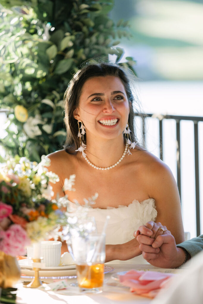 Candid portrait of bride smiling during toasts at her outdoor wedding reception wearing pearls and holding her husband's hand