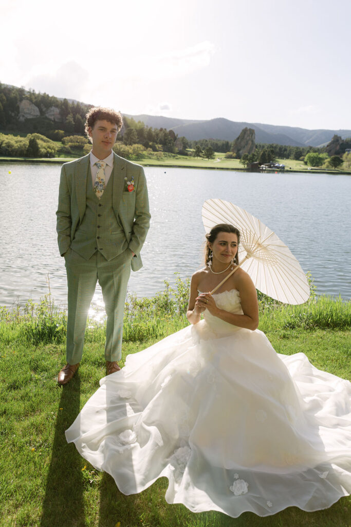 Bride in flowing white dress with parasol and groom in sage green suit in an editorial wedding photography pose at Perry Park Country Club Colorado wedding