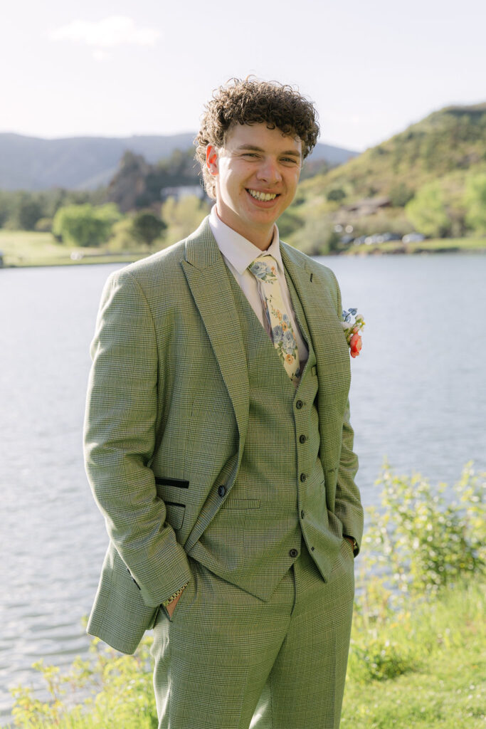 Groom in sage green three-piece suit with floral tie smiling by Lake Wauconda at Colorado Front Range wedding venue