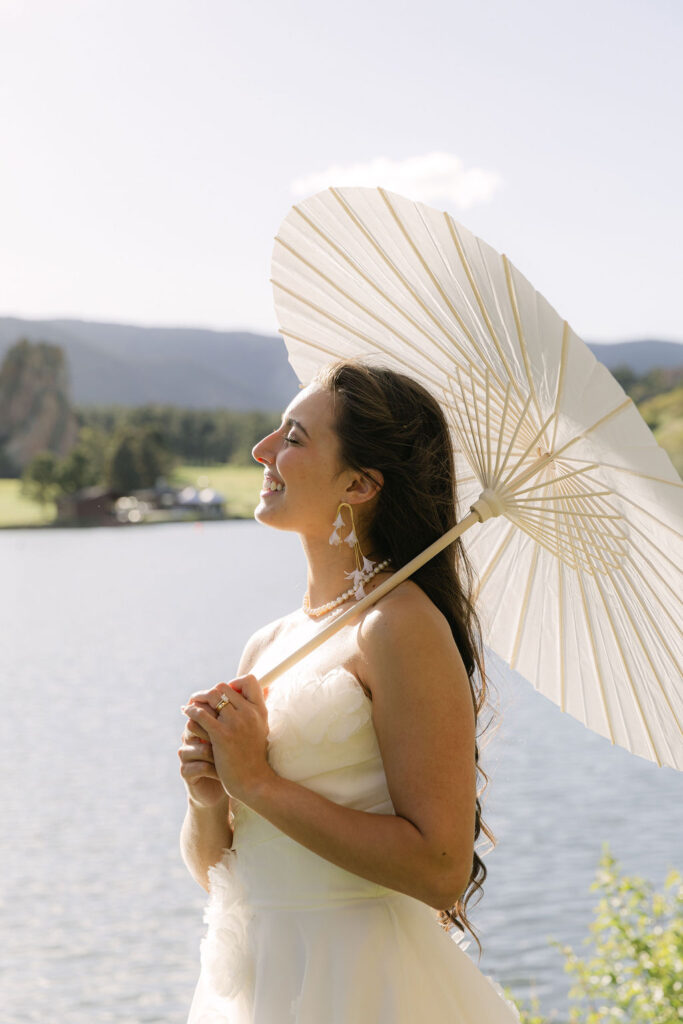 Bride smiling in the sunshine with a parasol by the lake at her Colorado wedding