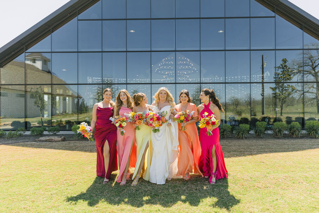 A joyful bridal party in colorful mismatched dresses poses in front of the glass-walled Camp Hosea reception building, perfect inspiration for a modern Texas wedding.