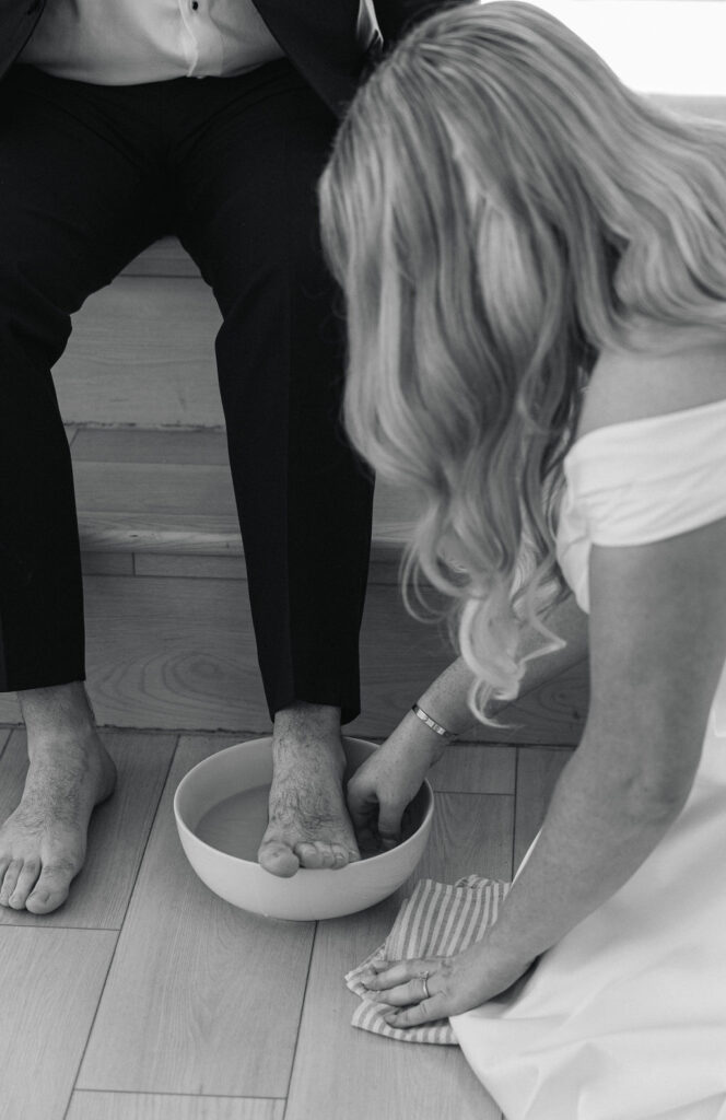 A black-and-white image of the bride washing the groom’s feet, a powerful and intentional wedding tradition rooted in faith, captured during this College Station wedding.