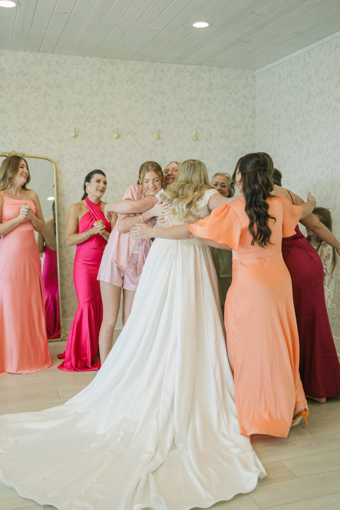 The bride, surrounded by bridesmaids in pink and coral dresses, shares a joyful moment while getting ready inside the Camp Hosea bridal suite.