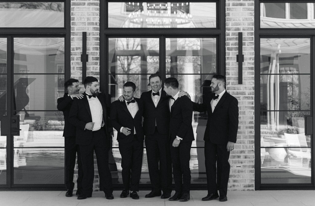 The groom poses with groomsmen in black tuxedos outside the reception hall, ready for this elegant Houston-area wedding.