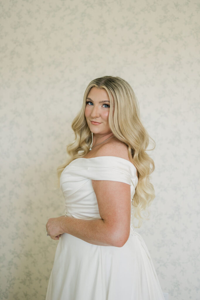 A bridal portrait with the bride smiling softly while standing in front of floral wallpaper, wearing an off-the-shoulder satin gown for her Camp Hosea wedding.