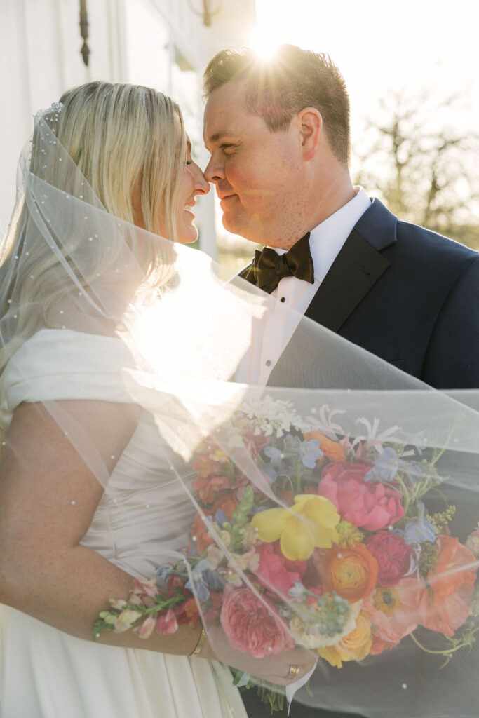 The couple share a kiss under the bride’s veil, with her bright bouquet glowing in the Texas sunlight during their Camp Hosea wedding portraits.