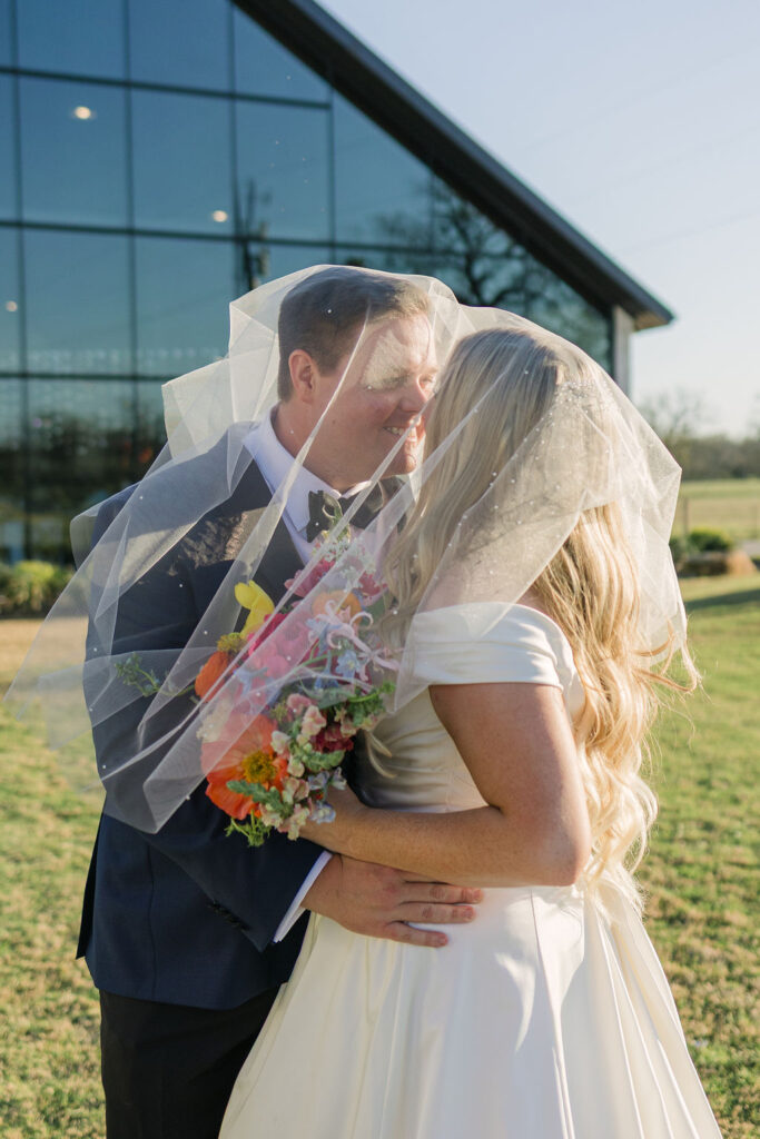 A soft veil-wrapped kiss between the newlyweds during portraits on the venue’s sunlit lawn.