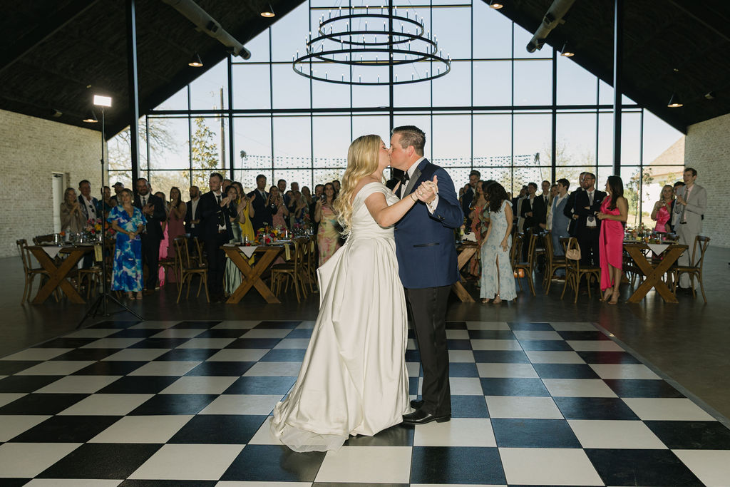 A photo of the bride and groom’s first dance on the black-and-white checkered floor surrounded by guests under the chandeliers of Camp Hosea.