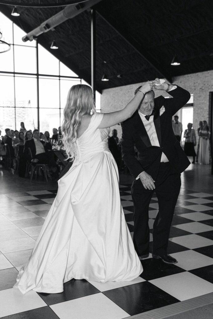 The bride twirls her dad in a romantic black-and-white photo during their first dance, surrounded by family and friends.
