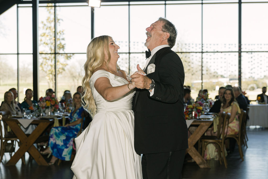 The bride laughs during her dance with her dad on the reception floor, her satin gown flowing around her in the light.