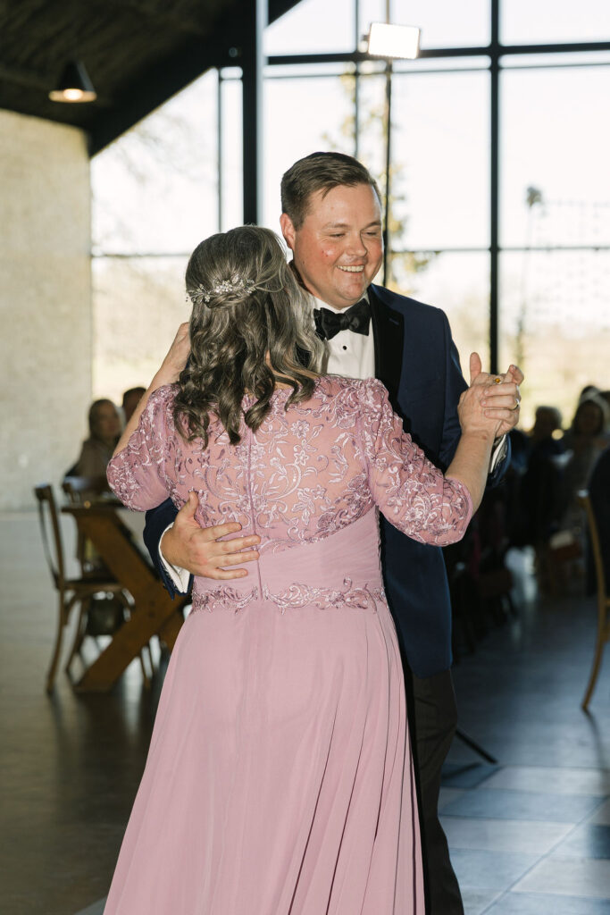 The groom shares a mother-son dance with his mom wearing a soft mauve gown, lit by windows at Camp Hosea’s reception space.