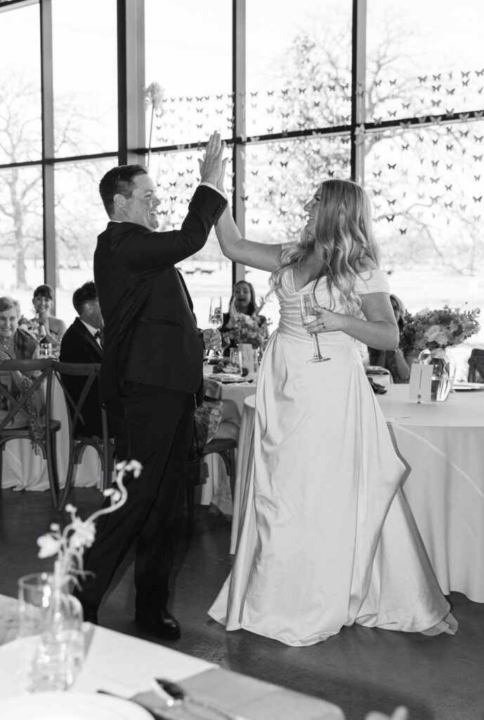 The groom high-fives the bride during toasts, celebrating under chandeliers in the Camp Hosea reception space.