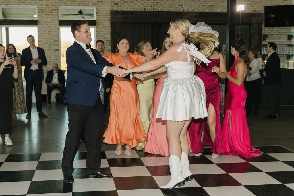 The bride and groom jump around while dancing with their friends, all dressed in bold spring colors inside the Camp Hosea reception hall.