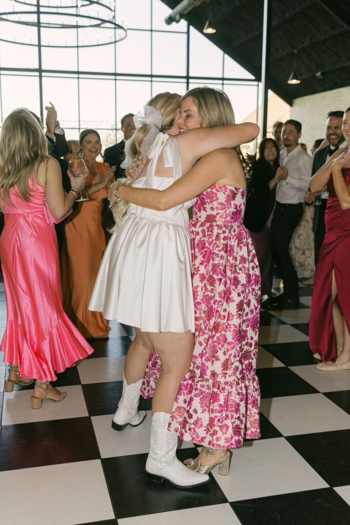 The bride hugs a guest wearing a pink floral dress while dancing on the checkered dance floor at Camp Hosea.
