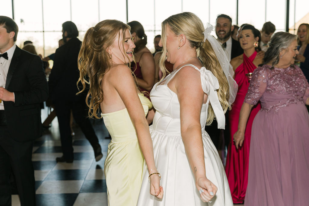 A joyful moment between the bride and a bridesmaid in lemon yellow as they laugh and dance at the reception.