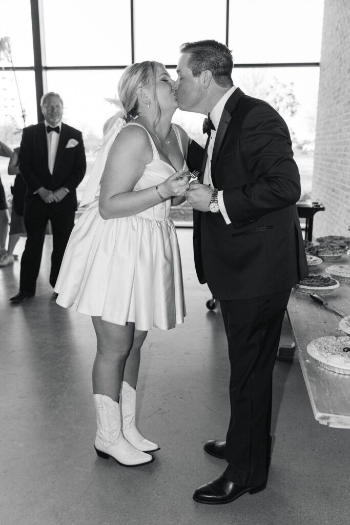 A black-and-white image of the couple sharing a kiss after cake cutting, highlighting joyful celebration moments at their modern Texas wedding.