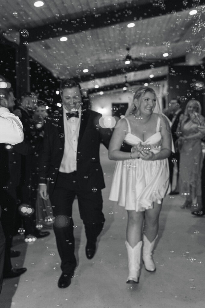A black-and-white image of the bride and groom walking through bubbles at their Camp Hosea wedding sendoff, surrounded by cheering guests.
