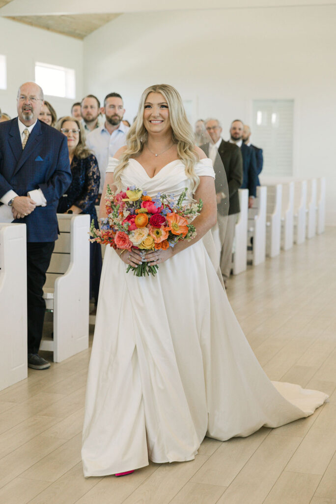 The bride smiles joyfully while walking down the aisle, holding a bright bouquet of ranunculus and peonies at this Camp Hosea wedding ceremony.