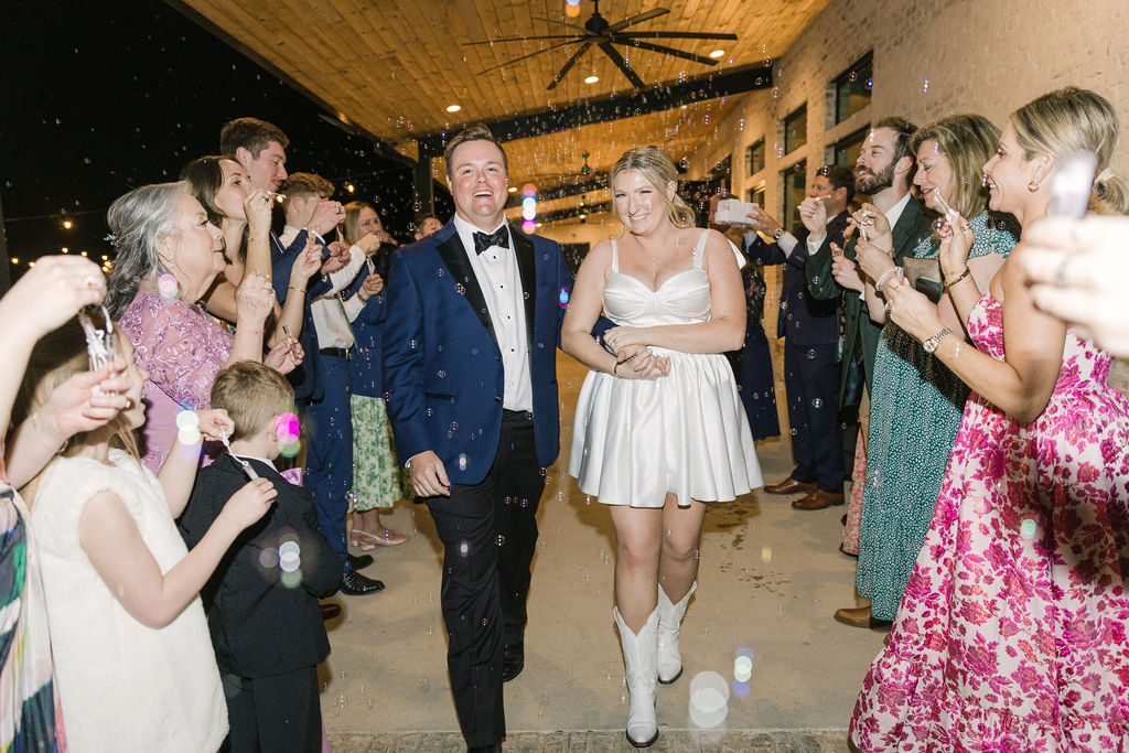 The newlyweds walk hand-in-hand down their bubble-filled sendoff tunnel, her short dress and boots perfect for their fun, modern exit.