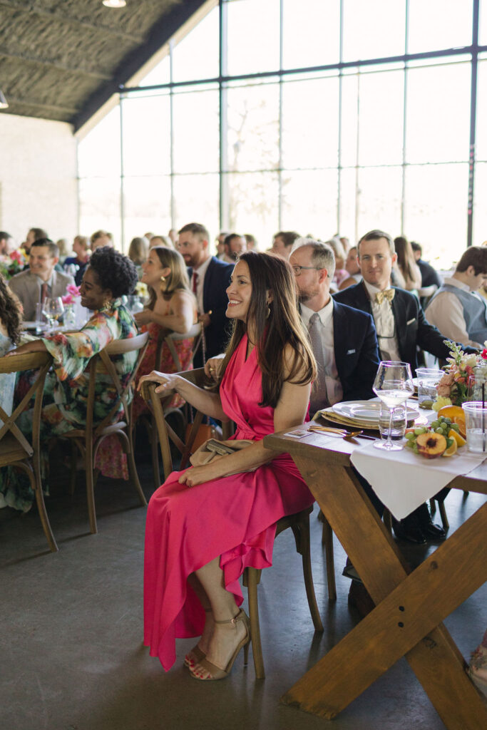 A guest in a hot pink dress sits at a wooden reception table, laughing and enjoying the colorful celebration.