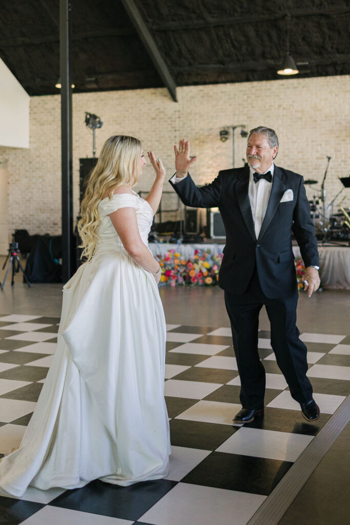 The bride high fives with her father during their heartfelt dance, surrounded by joyful guests and floral décor at Camp Hosea.
