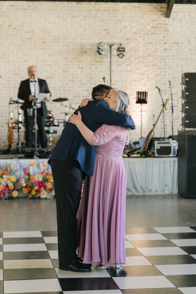 The groom shares a sweet dance with his mother on the black-and-white checkered dance floor under warm lighting.