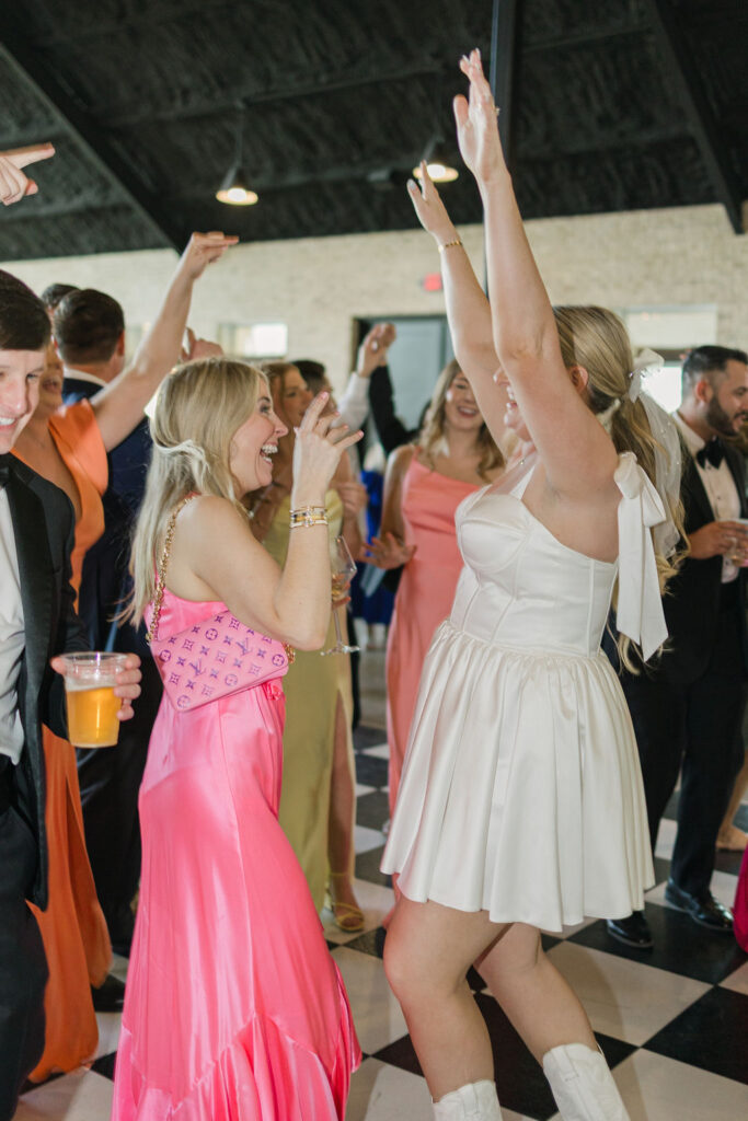 The bride in a short white dress and cowgirl boots dances with friends on the checkered floor at her Camp Hosea wedding reception.