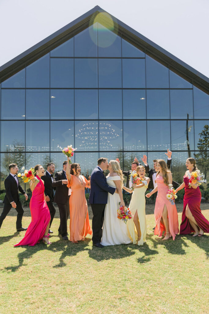 The bride and groom kiss in front of Camp Hosea’s dramatic glass chapel with their wedding party, dressed in vibrant spring tones.