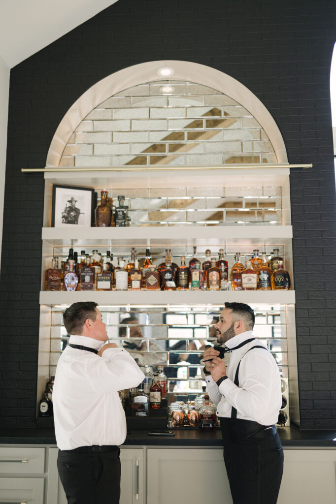 Two groomsmen adjust bow ties at a modern bar lined with whiskey bottles—part of the groom’s getting-ready moments at this Houston area wedding.