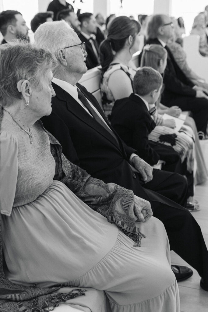A black-and-white photo of grandparents watching attentively from white pews inside this Houston-area wedding venue.