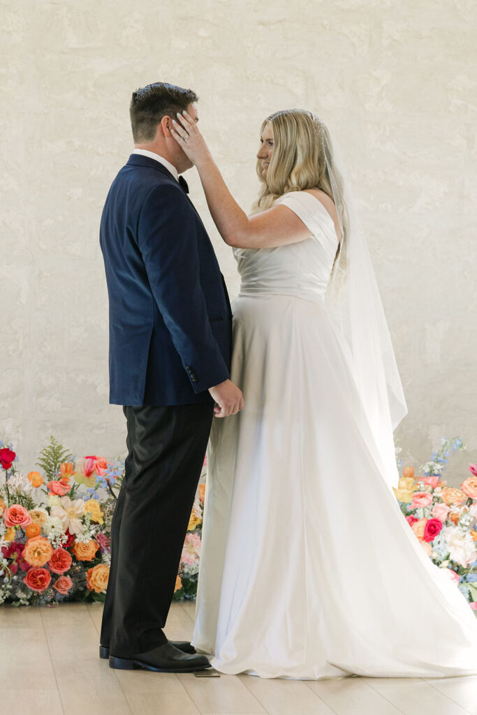 The bride lovingly places her hand on the groom’s cheek during the ceremony, standing on the altar steps surrounded by bright florals in the Camp Hosea chapel.