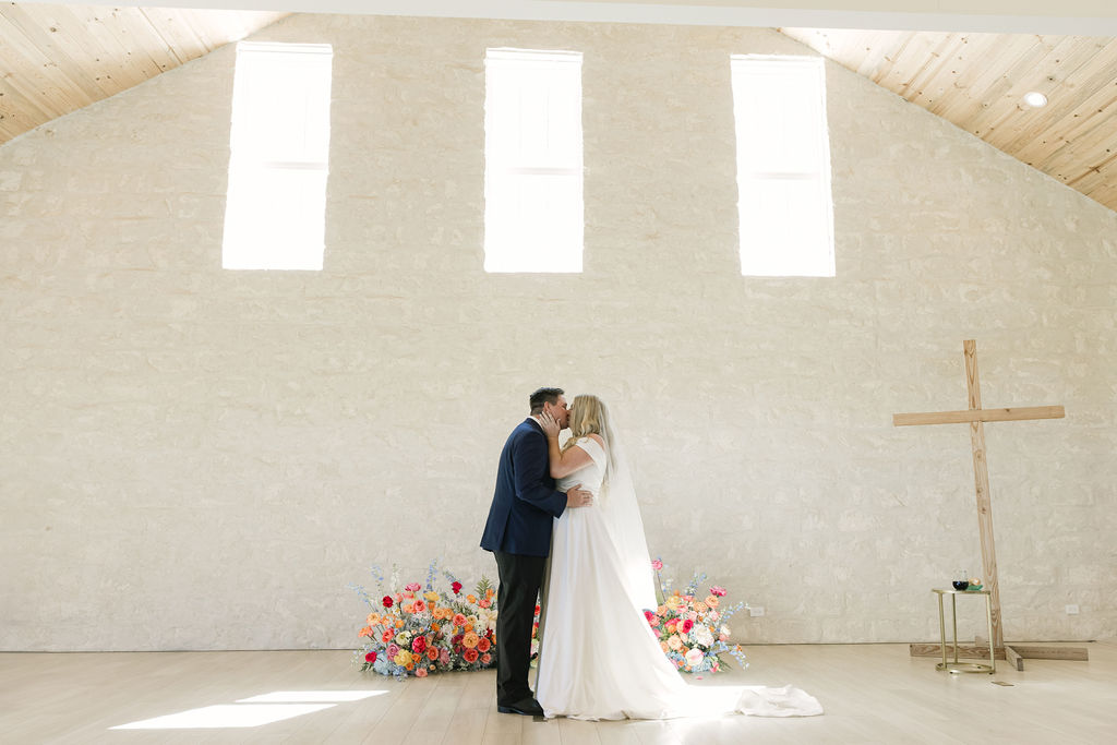 The couple kisses in front of the altar, framed by bright natural light and colorful spring florals in this modern Texas wedding venue.