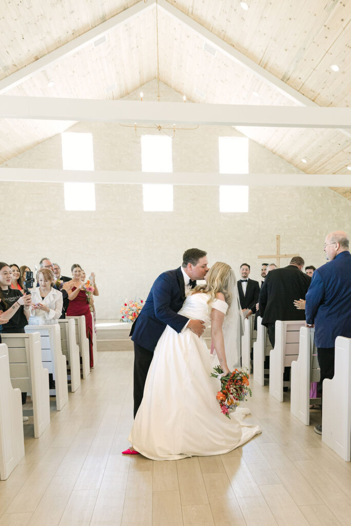 The groom dips the bride for a joyful kiss as they exit the aisle, surrounded by guests clapping and cheering in the Camp Hosea chapel.