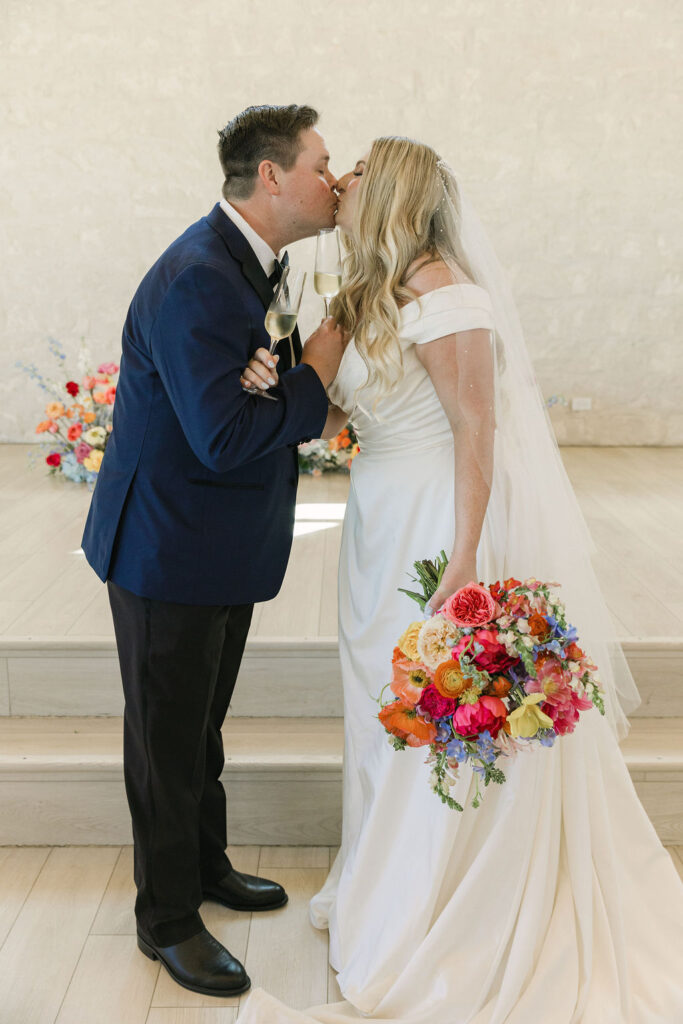 The newlyweds share a kiss after their ceremony, the bride’s colorful bouquet and soft veil catching the light inside this elegant Texas wedding venue.