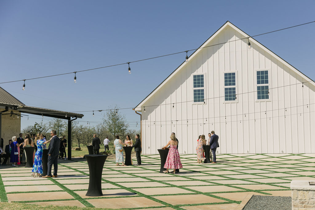 A wide shot of the cocktail lawn at Camp Hosea shows guests mingling in the courtyard under string lights.