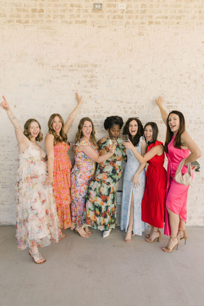 Guests pose for a group photo in colorful floral dresses outside the neutral stone backdrop of this College Station wedding venue.