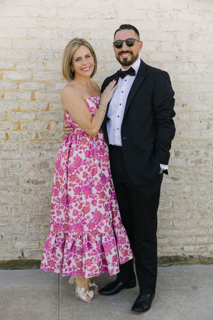 A couple poses together, the woman in a pink floral midi dress and the man in a black tuxedo—capturing wedding guest style at this elegant Texas wedding.