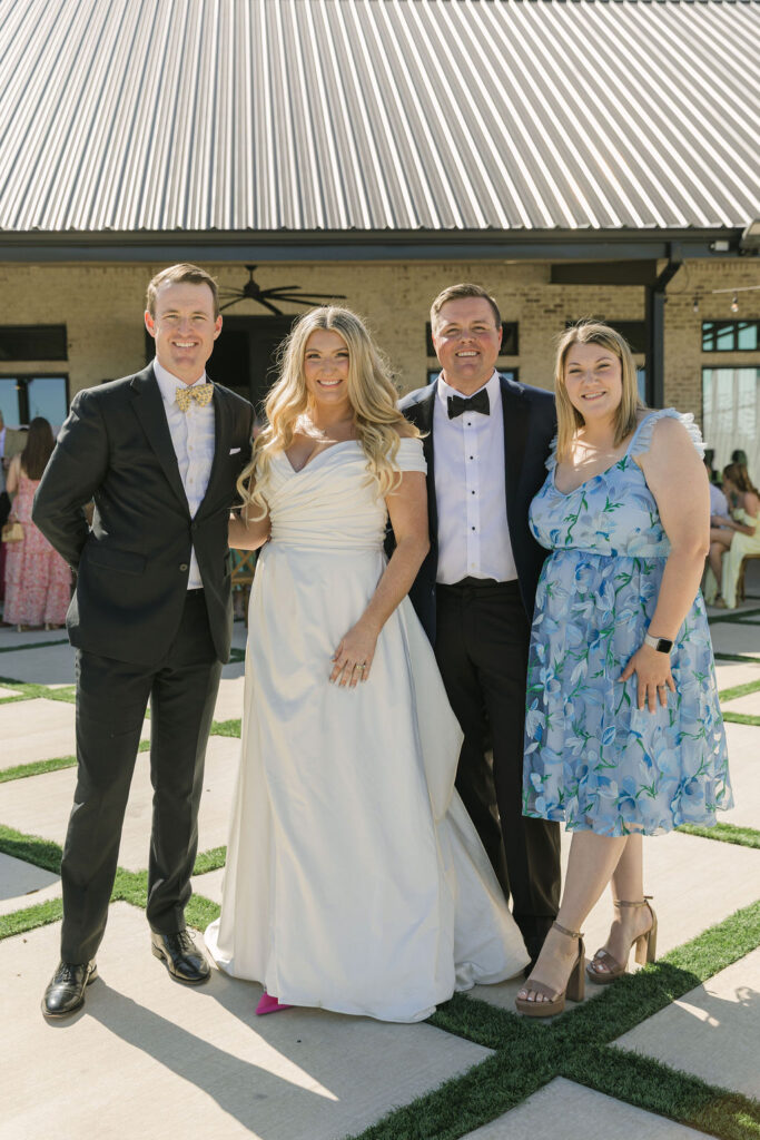 The bride and groom pose with family members outside the reception building, the bride glowing in her off-shoulder gown.