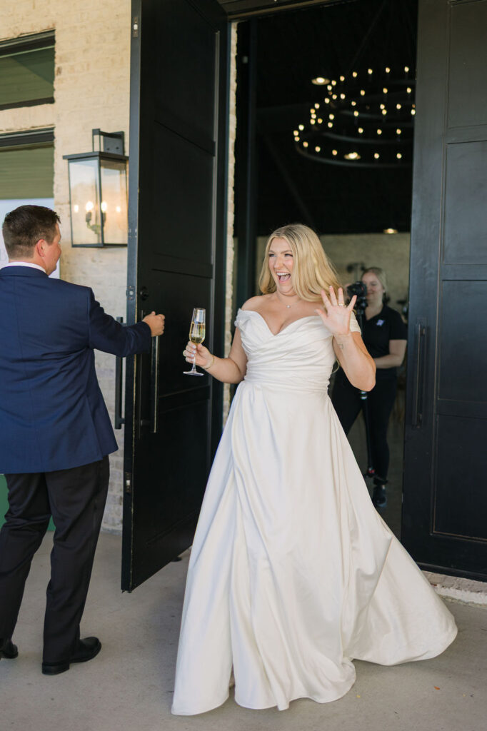 The bride raises her arms in celebration while exiting the venue’s grand black doors, framed by springtime light and modern architectural details.